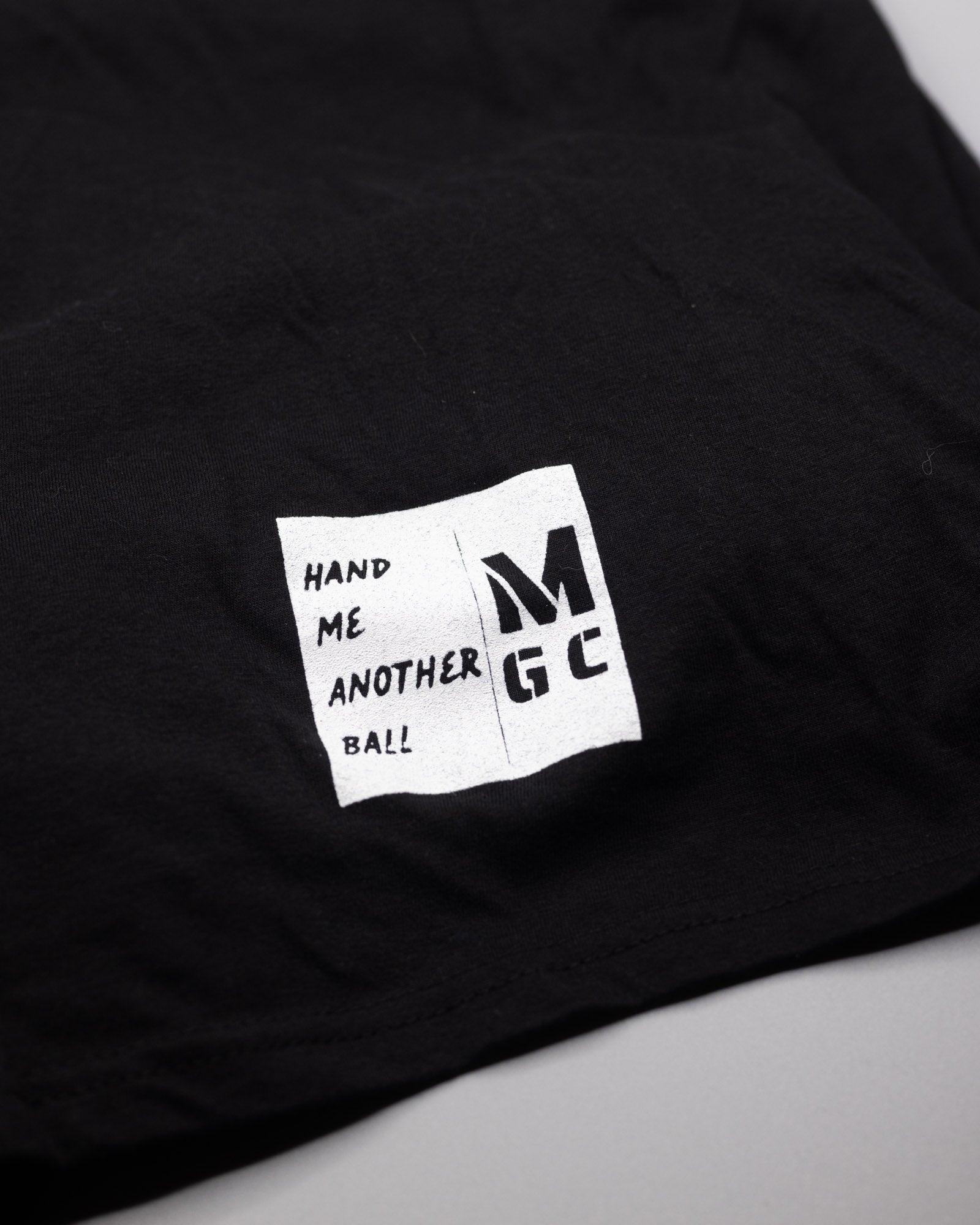 This is the MLGN "Built" black, ringspun, t-shirt with the Mully logo centered on the shirt with the words MLGN GOLF arched over the logo and the word COMPANY and 2024 underneath the logo.
On the bottom right corner of the shirt there is a small white coloured screened on feature tag that reads "Hand Me Another Ball" in black writing. The letters MGC are beside the Hand Me Another Ball saying.